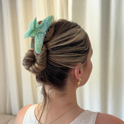 Blonde woman with a bun held by a seafoam green starfish-shaped hair claw clip, wearing gold earrings.