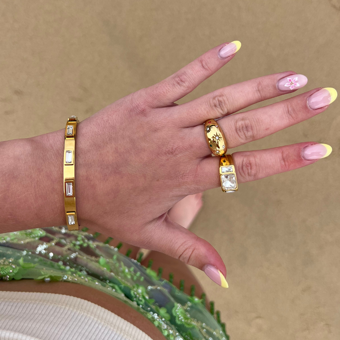 Gold Coconut Lane Stargaze Signet Ring on a hand with yellow-tipped nails, above sand and a green beaded skirt.