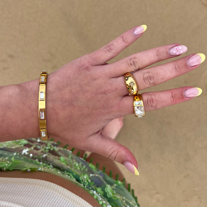 Gold Coconut Lane Stargaze Signet Ring on a hand with yellow-tipped nails, above sand and a green beaded skirt.