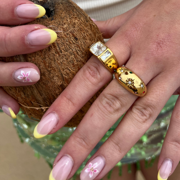 Hands with yellow-tipped floral nails and a gold Coconut Lane Stargaze signet ring hold a coconut.