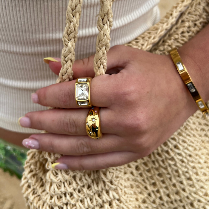Hand with yellow-accented nails wears a gold Stargaze signet ring, holding a tan woven bag.