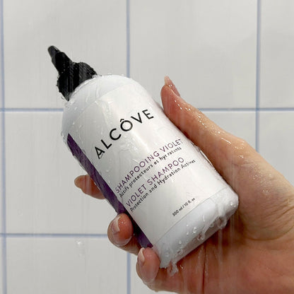 Hand holding a bottle of Violet Shampoo - Alcôve Hair in a bathroom