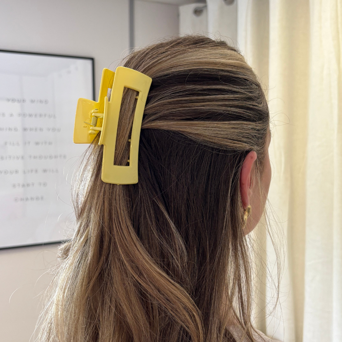 Blonde woman wears a yellow square claw clip, holding her hair back, standing indoors near a window.